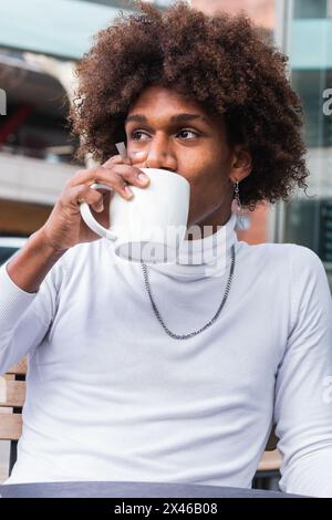 Relaxed young black guy with Afro hairstyle in stylish clothes drinking coffee and looking away while sitting on cafe terrace in city Stock Photo