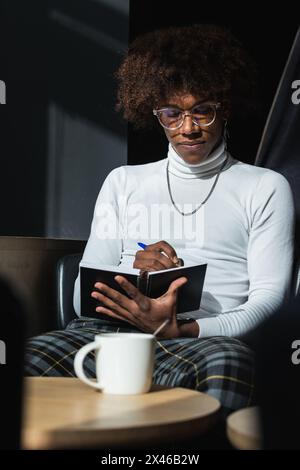 Confident young African American male student with curly hair in stylish outfit and eyeglasses taking notes in copybook while preparing for exam in mo Stock Photo