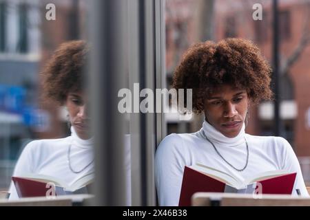 Calm young African American guy with curly hair in trendy outfit resting on cafe terrace and reading interesting book Stock Photo