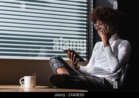 Young ethnic male freelancer in stylish clothes and eyeglasses taking notes in planner and using smartphone while working remotely at table with cup o Stock Photo