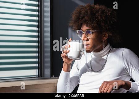 Relaxed young black guy with Afro hairstyle in stylish clothes drinking coffee and looking away while sitting on cafe terrace in city Stock Photo