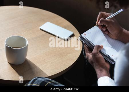 Crop young African American male student with curly hair in stylish outfit and eyeglasses taking notes in copybook while preparing for exam in modern Stock Photo