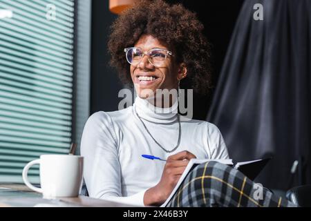 Cheerful young African American male student with curly hair in stylish outfit and eyeglasses taking notes in copybook while preparing for exam in mod Stock Photo