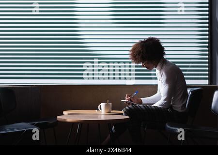 Side view of concentrated young African American male student with curly hair in casual clothes taking notes in copybook while doing homework assignme Stock Photo