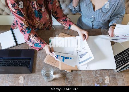 From above crop female coworkers gathering at desk with laptops and analyzing diagrams while working together in modern workspace Stock Photo