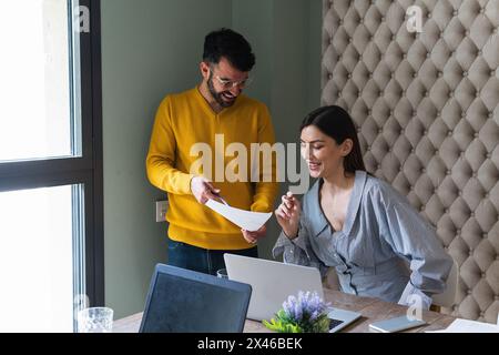 Happy young coworkers gathering and desk with laptops and discussing documents details in light modern workplace Stock Photo