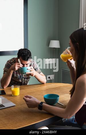 Cheerful young friends wearing casual outfits enjoying tasty lunch and chatting while sitting at dining table with opened netbook in modern apartment Stock Photo