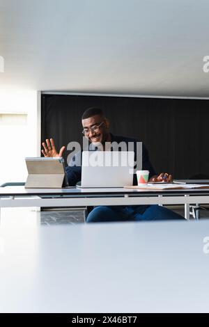 Positive African American adult male in formal clothes and eyeglasses sitting at table while having video call on tablet near netbook and documents wi Stock Photo