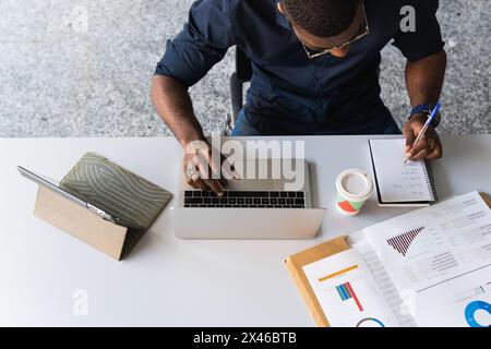 From above black adult male in formal clothes sitting at table while taking notes in notebook from papers near netbook in bright workplace Stock Photo