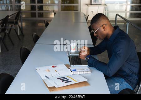 Side view of African American concentrated male in eyeglasses and formal clothes sitting in chair at table while using phone and netbook near document Stock Photo