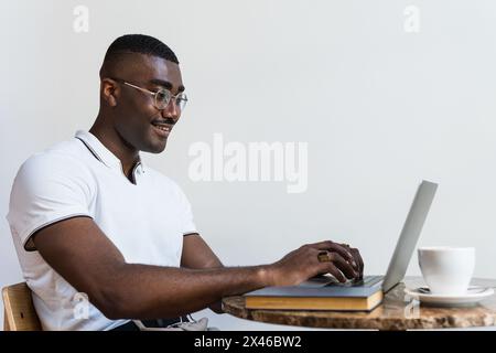 Happy black adult male in eyeglasses and casual clothes sitting in chair at table in light cafeteria and browsing on netbook near cup of coffee Stock Photo