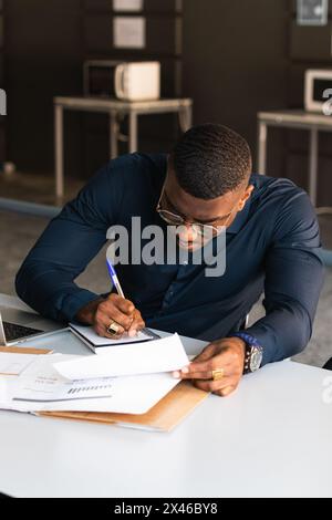 Serious black adult male in formal clothes and eyeglasses sitting at table while taking notes in notebook from papers near netbook in bright workplace Stock Photo