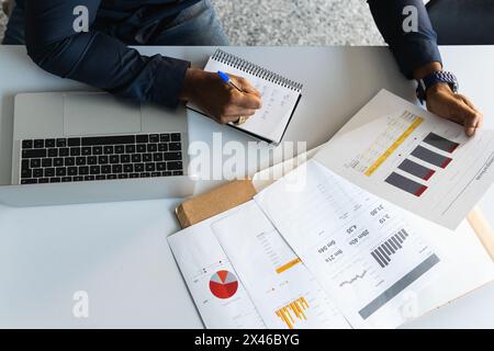 From above black adult male in formal clothes sitting at table while taking notes in notebook from papers near netbook in bright workplace Stock Photo