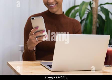 Cropped unrecognizable happy African American female freelancer browsing smartphone and smiling while sitting at table with laptop in cafe and working Stock Photo