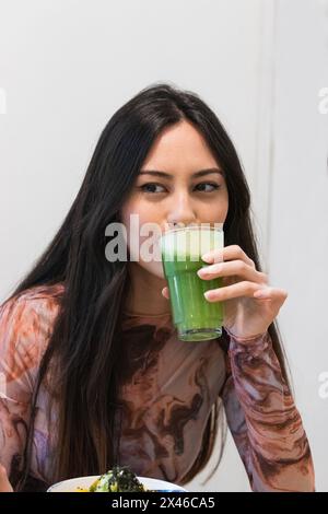 Delighted female sitting in restaurant and drinking refreshing green smoothie while enjoying weekend and looking away Stock Photo