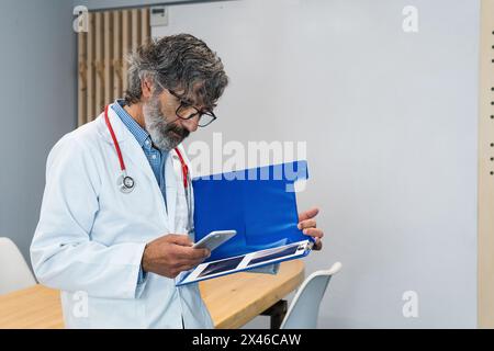 Focused elderly male medic in uniform and with x rays in folder standing in hospital and browsing mobile phone Stock Photo