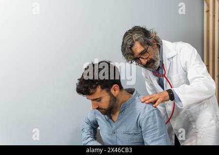 Elderly male doctor examining patient with stethoscope during appointment in hospital against white wall Stock Photo