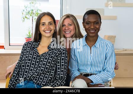 Delighted multiracial female colleagues looking at camera and smiling while sitting in workplace together Stock Photo