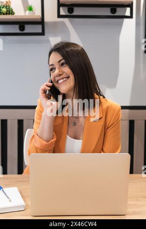 Smiling female freelancer sitting at table with laptop and using a phone Stock Photo