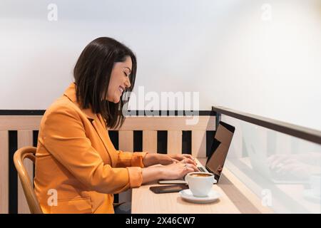 Side view of happy female freelancer browsing netbook while sitting at table with cup of coffee and working remotely in cafe Stock Photo