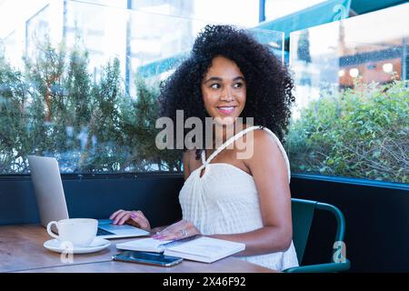 Pleasant pregnant African American female freelancer working on netbook at table with cup of coffee notepad and smartphone Stock Photo