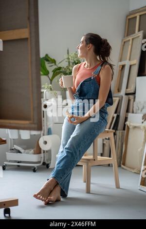 Artist lunch in home studio, freelancer working break. Young female painter relaxes with coffee cup Stock Photo