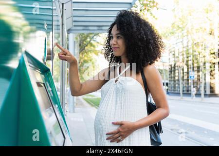 Pregnant African American female touching tummy while searching information on display for navigation on city street Stock Photo