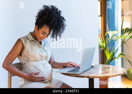 Positive pregnant African American female with curly hair touching tummy while typing on netbook at table Stock Photo
