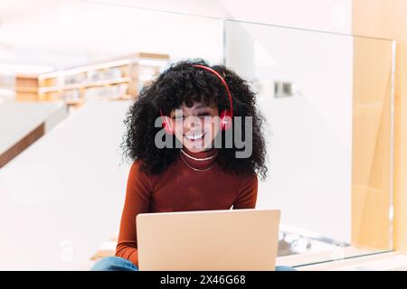 Black curly haired cheerful female with headphones while sitting with laptop in modern library Stock Photo
