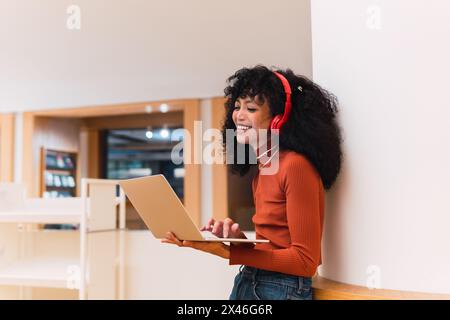 Black curly haired cheerful female with headphones while standing with laptop in modern library leaning on wall Stock Photo