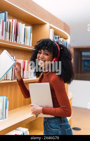 Side view of smiling black female with headphones and laptop looking at camera while taking book from bookcase in library Stock Photo