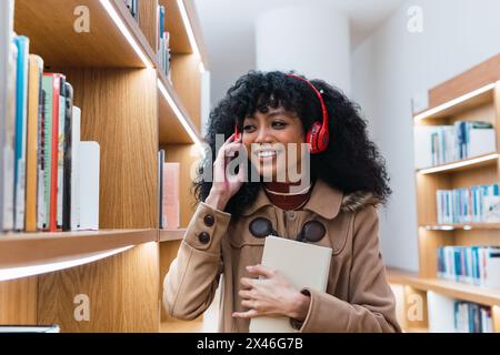 Happy African American female student in headphones talking on mobile phone and holding book near bookcase in university library Stock Photo