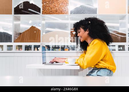 Side view of young cheerful African American female freelancer sitting at table with cup of coffee and typing on laptop Stock Photo