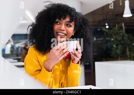Content young ethnic female with long Afro hair smiling and drinking cup of coffee in light cafe Stock Photo