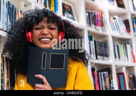 Stylish happy young ethnic female student with long dark curly hair in trendy outfit and headphones smiling while holding with book in modern library Stock Photo