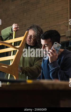 Busy male joiner measuring chair detail while colleague talking on mobile phone and writing order on paper Stock Photo