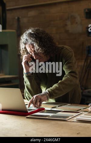 Concentrated male carpenter having phone call on mobile phone while typing order on netbook during work in joinery Stock Photo