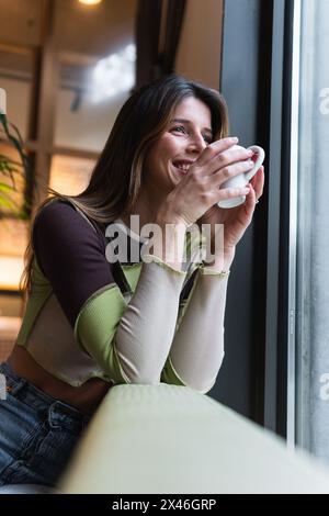 Young content female with cup of hot drink leaning on hand and looking away against window in cafeteria Stock Photo