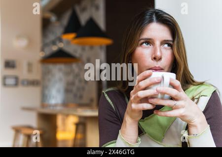 Young dreamy female blowing on hot drink in cup while looking up in cafeteria on blurred background Stock Photo