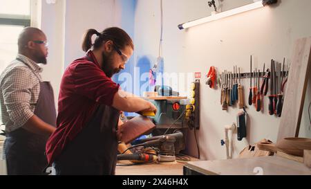 Carpenter doing last touches on piece of wood before using it for furniture assembly, sanding it with sandpaper. Woodworking expert doing woodworking process on lumber block, fixing damages, camera B Stock Photo
