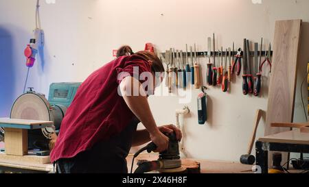 Carpenter wearing safety glasses giving clipboard with blueprints to assistant, using angle grinder on wood. Man with protective goggles using orbital sander gear after consulting schematics, camera A Stock Photo