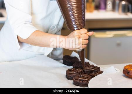 Crop unrecognizable female baker in mask squeezing chocolate cream from pastry bag on tasty vegan sponge cakes while cooking desserts in kitchen of ba Stock Photo