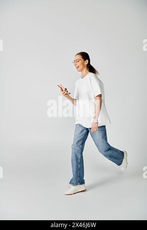 A young woman in a white t-shirt and glasses walks while focused on her cellphone screen. Stock Photo