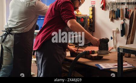 Craftsperson at work consulting blueprint and using orbital sander with sandpaper on lumber. Woodworking expert in carpentry shop uses angle grinder on wood after checking schematics, camera A Stock Photo