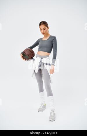 A sporty young woman gracefully holds a basketball in her right hand, dressed in active wear against a grey background. Stock Photo