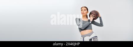 A sporty young woman elegantly holds a basketball in her right hand against a grey background. Stock Photo