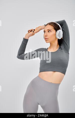 A sporty young woman in active wear strikes a pose as she listens to music through headphones on a grey background. Stock Photo