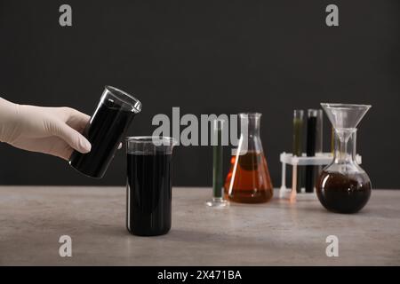 Woman pouring black crude oil into beaker at grey table against dark background, closeup Stock Photo
