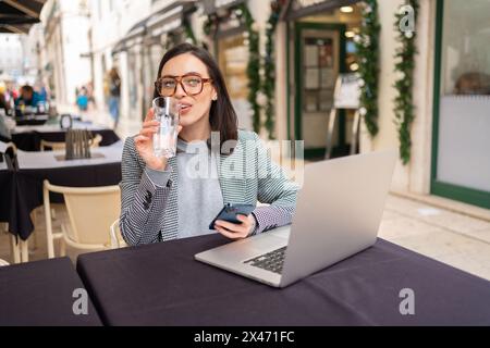 Portrait of attractive delighted charming smiling woman freelancer or student wearing glasses, working in cafe on laptop, holding mobile phone in hands, drinking water from glass. Stock Photo