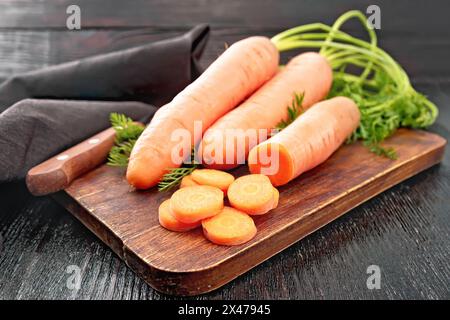 Fresh whole carrots with haulm and slices, knife and brown napkin on a background of a dark wooden board Stock Photo